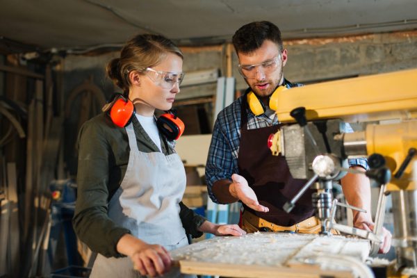 Listening to mentor with attention Waist-up portrait of attractive apprentice standing at drill press and listening to her male mentor with attention, he explaining her operating principle of machine